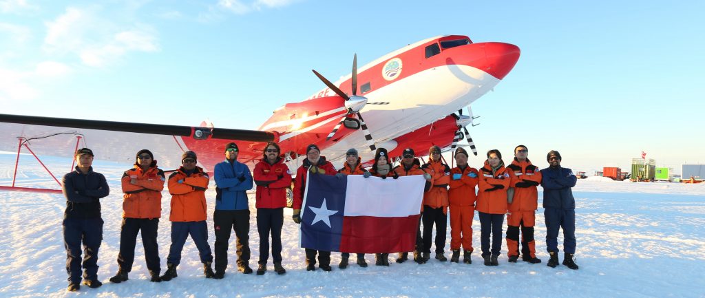 A group of Antarctic researchers poses in front of a propeller plane holding a flag of Texas between them.