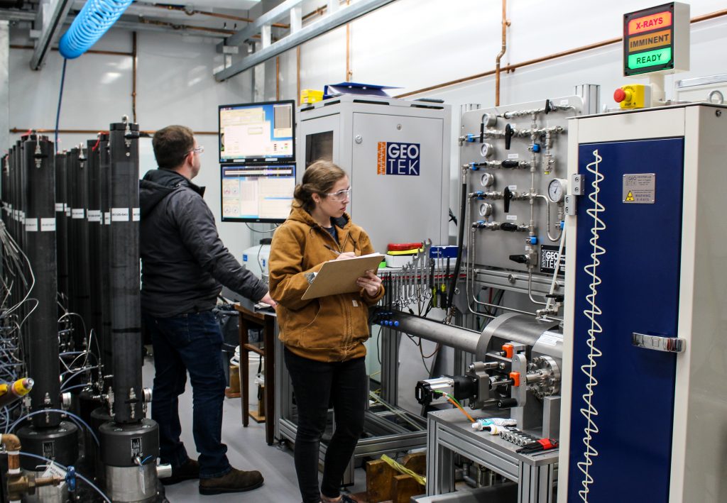 Photo of Addison and Josh in the lab both wearing thick cold weather coats and protective gear. Josh is working on a terminal and Addison is holding a clipboard and checking off readings. Behind them are two vertically stacked lines of 8 foot tall grey dark pressure cores.