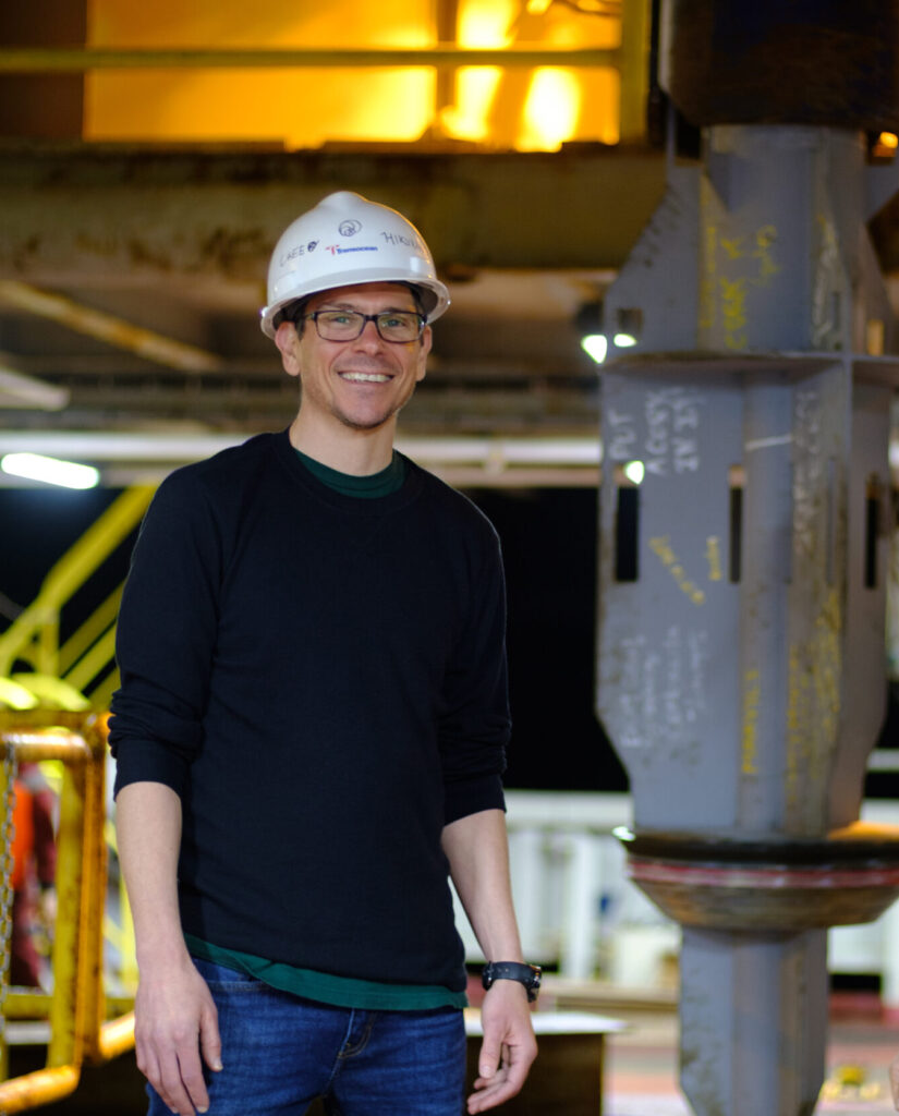 Demian with hard hat on the drilling ship, posing for the camera next to one of the borehole observatories