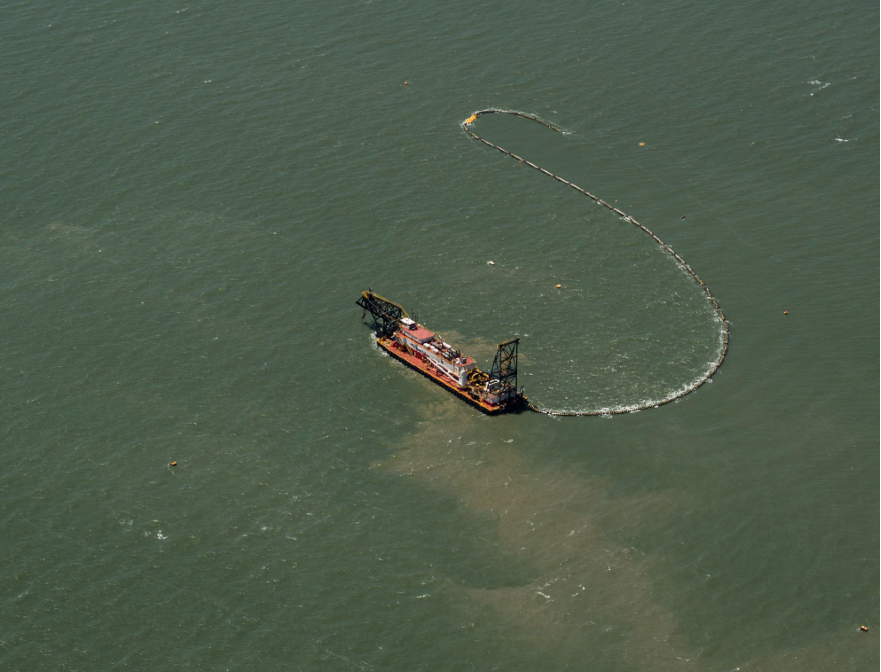 An aerial view of a dredging vessel at sea. Trails of sand are visible in vessel's wake.