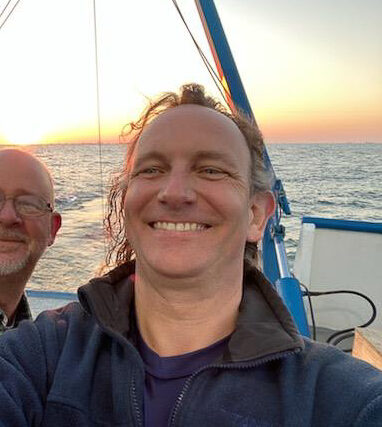 Picture of Sean taking a selfie at sea. The sun is shining on the sea behind him