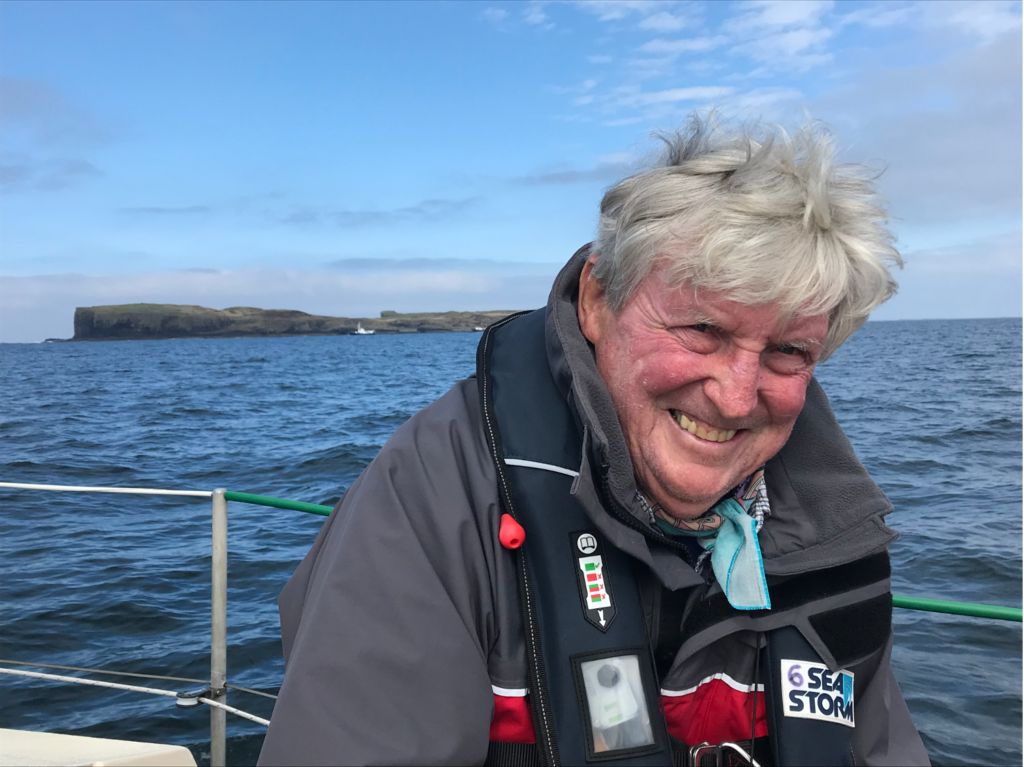 Ian Dalziel at sea wearing a storm jacket and life vest. A rugged windswept island fills the horizon. Dalziel is smiling at the camera.