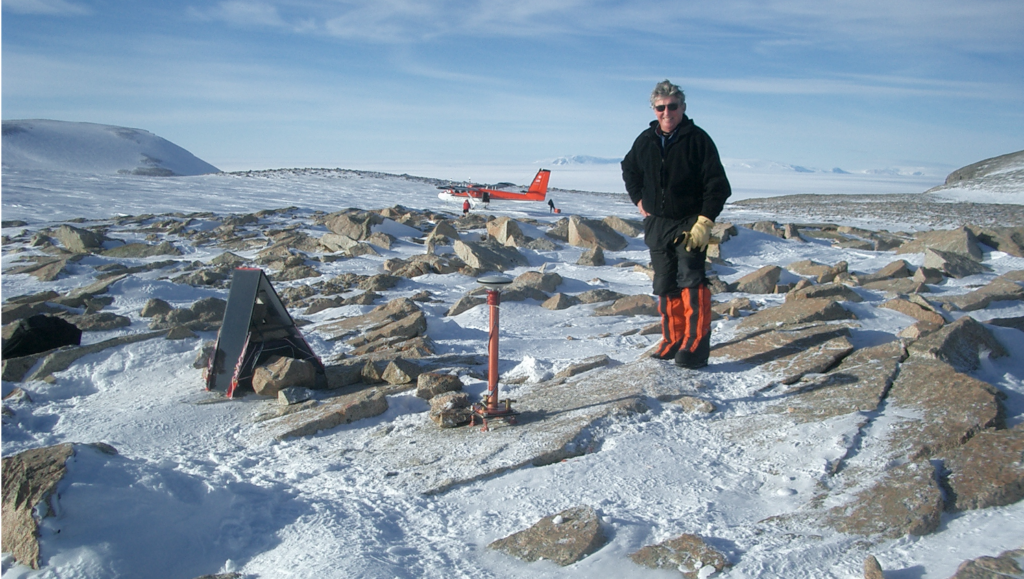 A picture showing Ian Dalziel standing among snow and rocks with a deployed sensor ahead of him. In the background is a landed Twin Otter aircraft.
