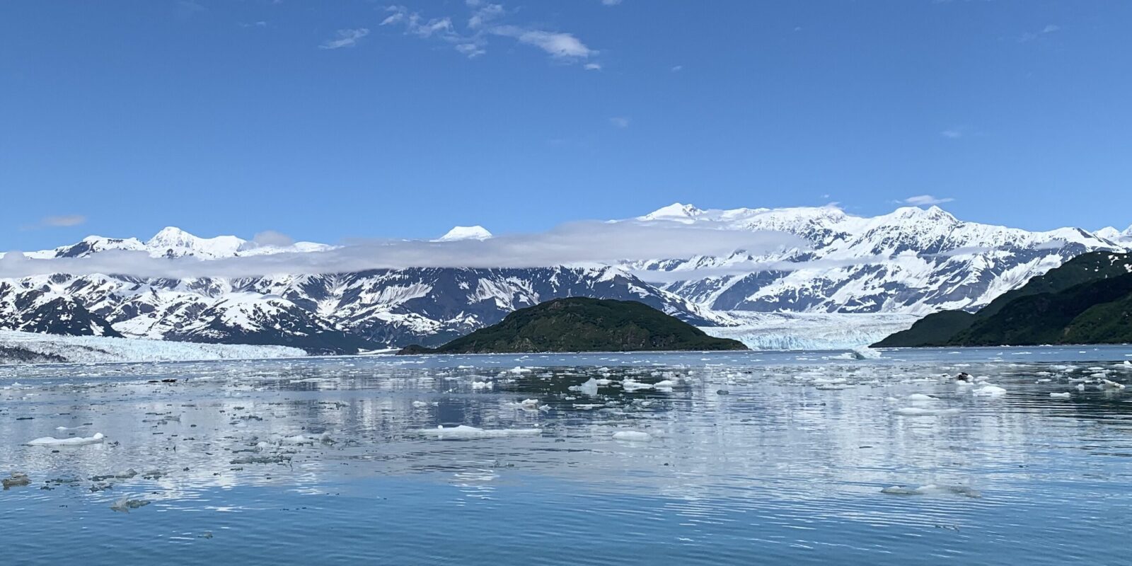 Blue skies reflected on a glacial fjord. The glacier and ice covered mountains are visible in the distance.
