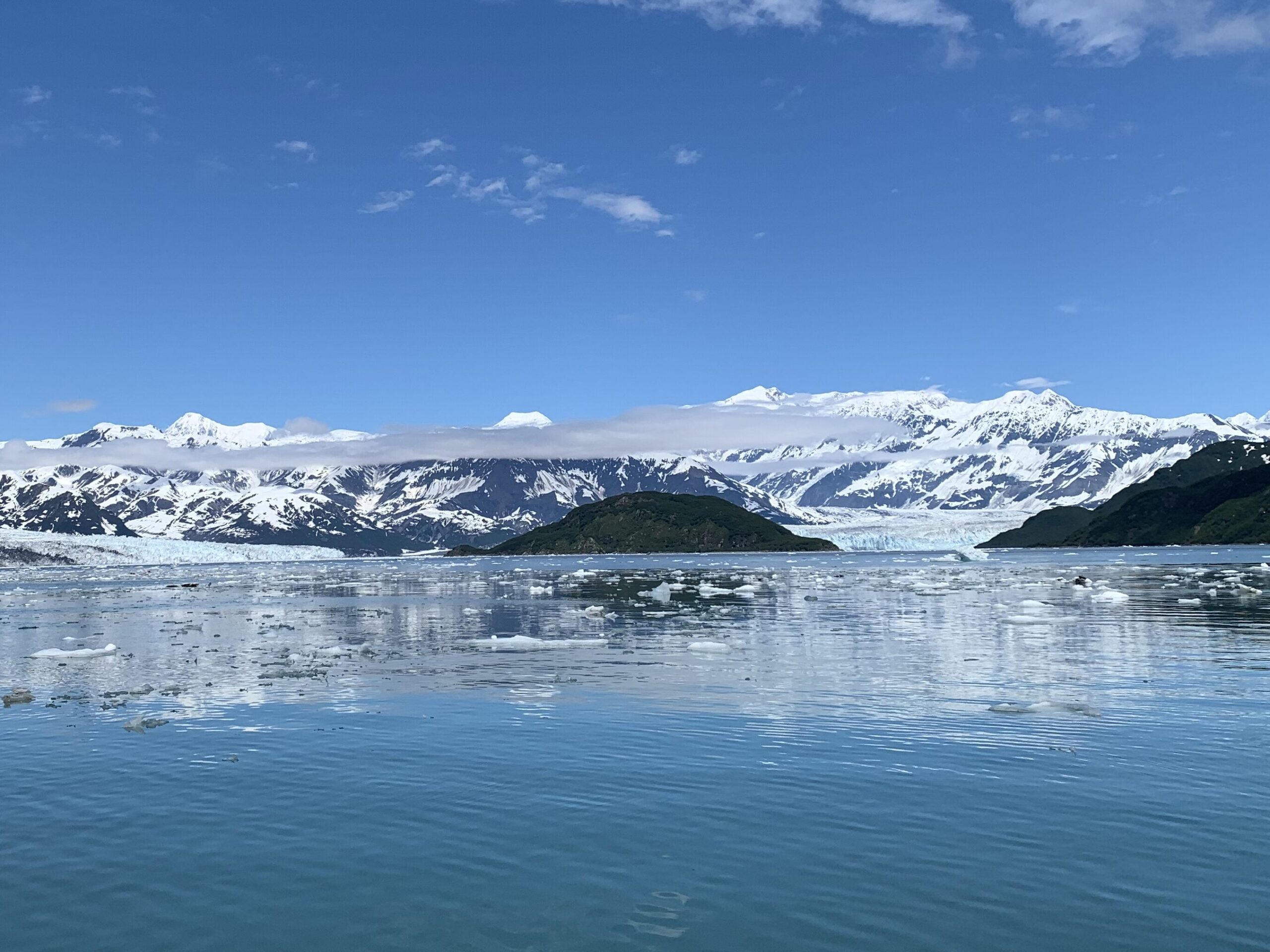 Blue skies reflected on a glacial fjord. The glacier and ice covered mountains are visible in the distance.