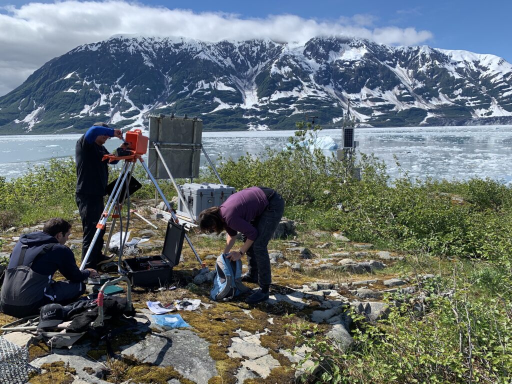 Researchers installing an instrument on land. Behind them is a glacial landscape.