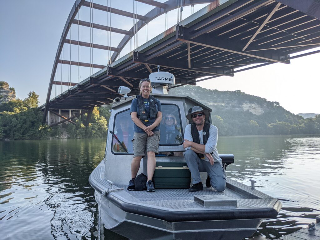 Boat with scientists standing on deck and inside, floats beneath Austin's bridge.