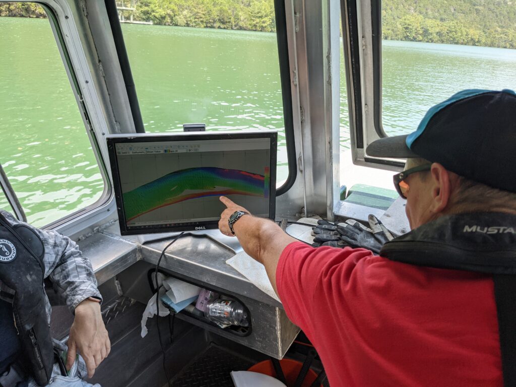 A scientist points at a scree showing colored sonar bathymetry