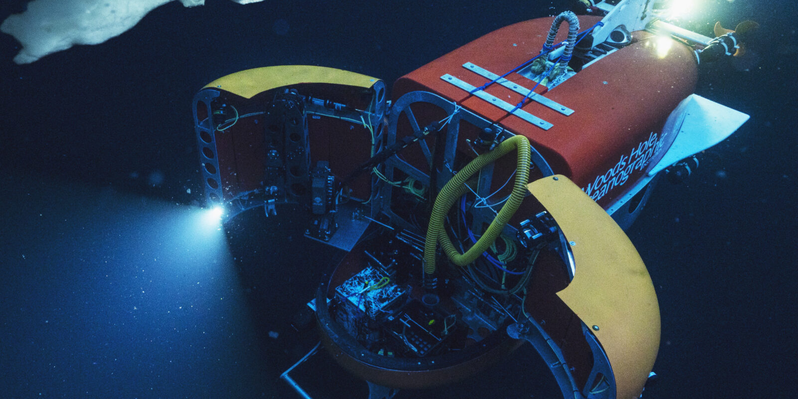 The robot in action underwater. The lights from its open payload bay doors light up ice underwater