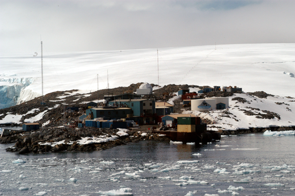 Low buildings perched on the shore with ice covered hills behind.