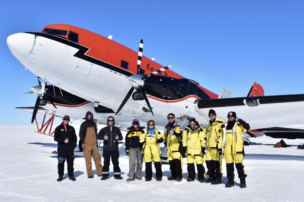 A group of nine scientists dressed for polar research pose in front of a propeller plane on the ice