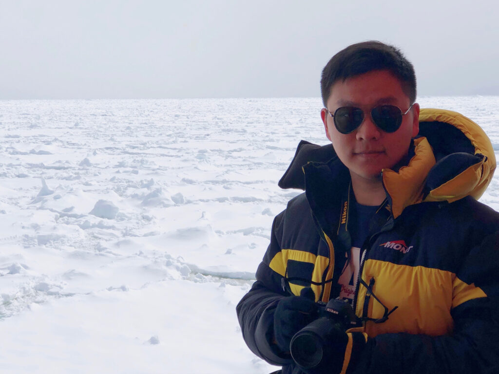 Photo of Shuai Yan in Antarctica. He is holding a camera and behind him is a flat landscape of ice.