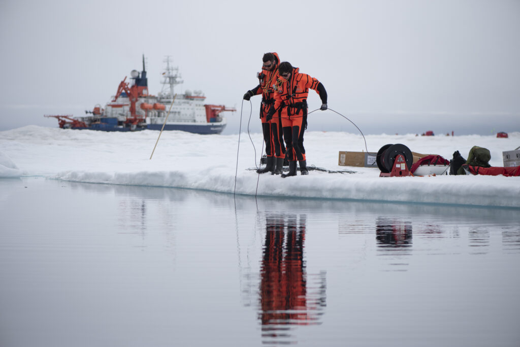 Two scientists in Arctic gear lower an instrument into the sea from the edge of a low iceshelf. Begin them an ice breaker ship is visible.