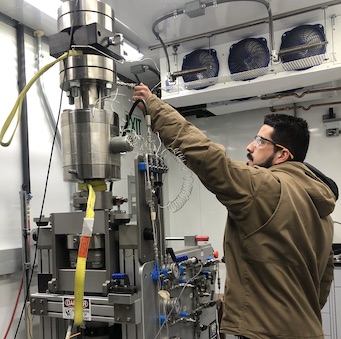 Picture of Alejandro working in the lab and adjusting a tall piece of equipment. The lab is climate controlled so he's wearing a thick winter coat.