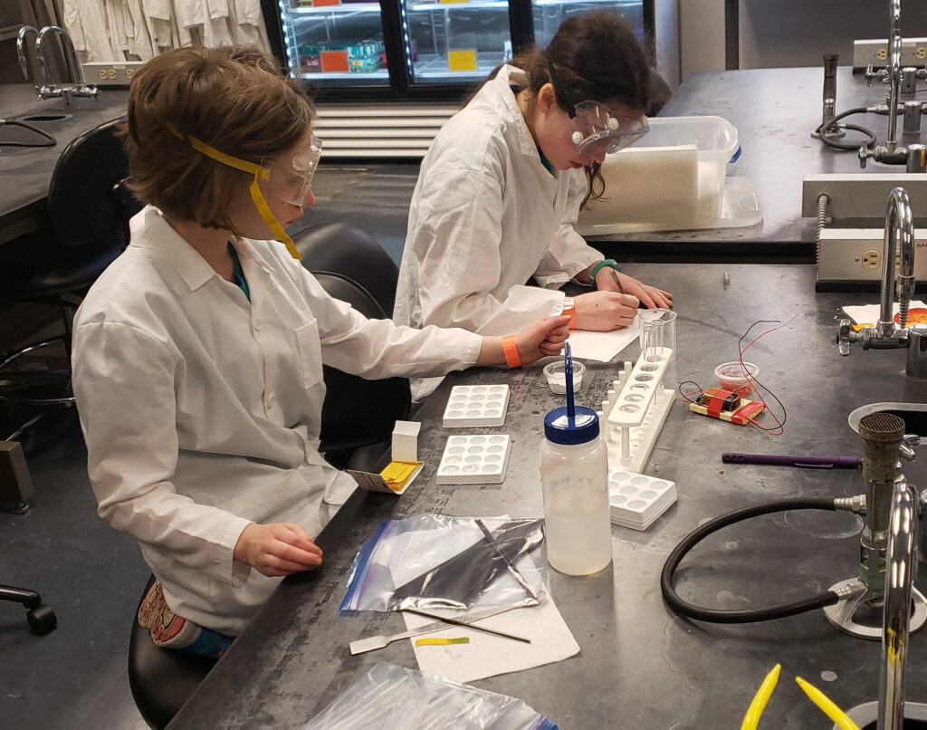 Two young students wearing lab coats and PPE focused on a chemistry task in a lab