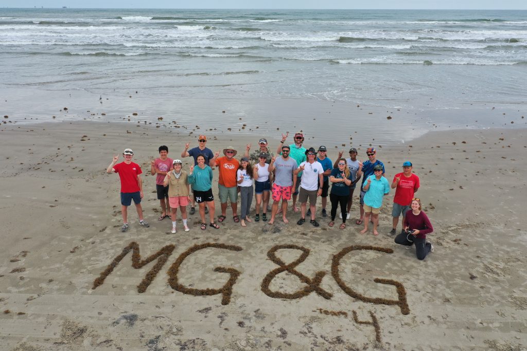 Group photo of the field class on a beach in front of the letters MG&G traced out in the sand. The photo is taken from above (drone).