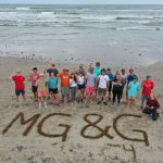 Group photo of the field class on a beach in front of the letters MG&G traced out in the sand. The photo is taken from above (drone).