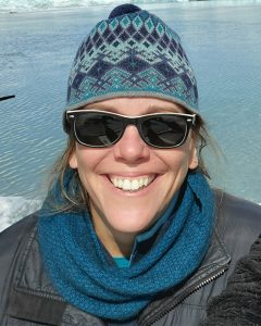 Portrait photo of Marcy with sunglasses, wooly hat and scarf near ice and water.