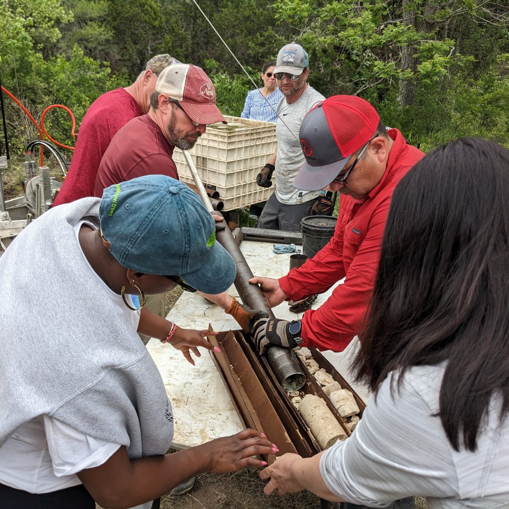 People wearing hats gathered round a table as a long metal tube is emptied of its rocky contents.