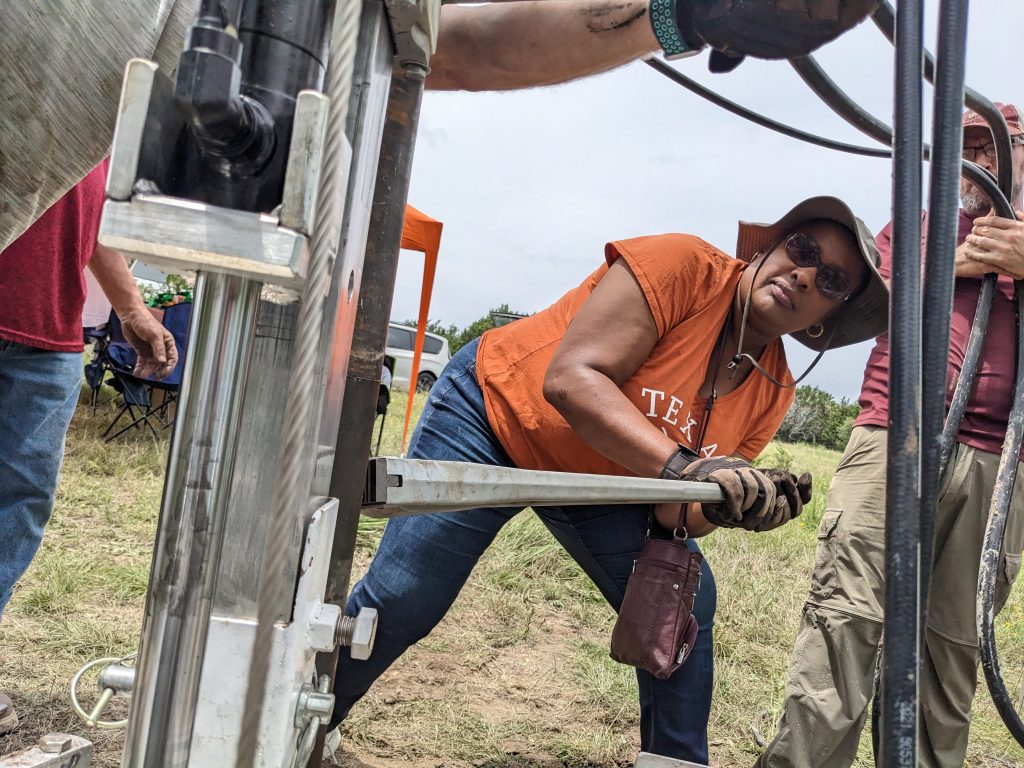 Rosalind straining against a large wrench that's gripping a vertical metal column. She is in a field and other people are visible around her.