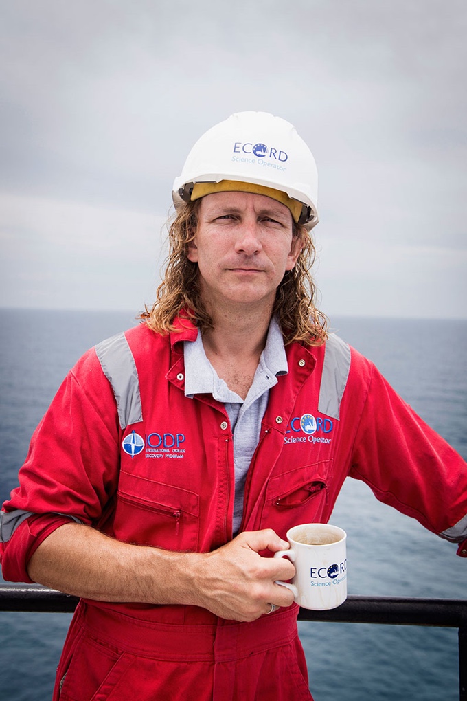 Portrait photo of Sean in hero pose. He's wearing a red boiler suit and hard hat and has the ocean behind him. He's holding a coffee mug with the ECORD logo.