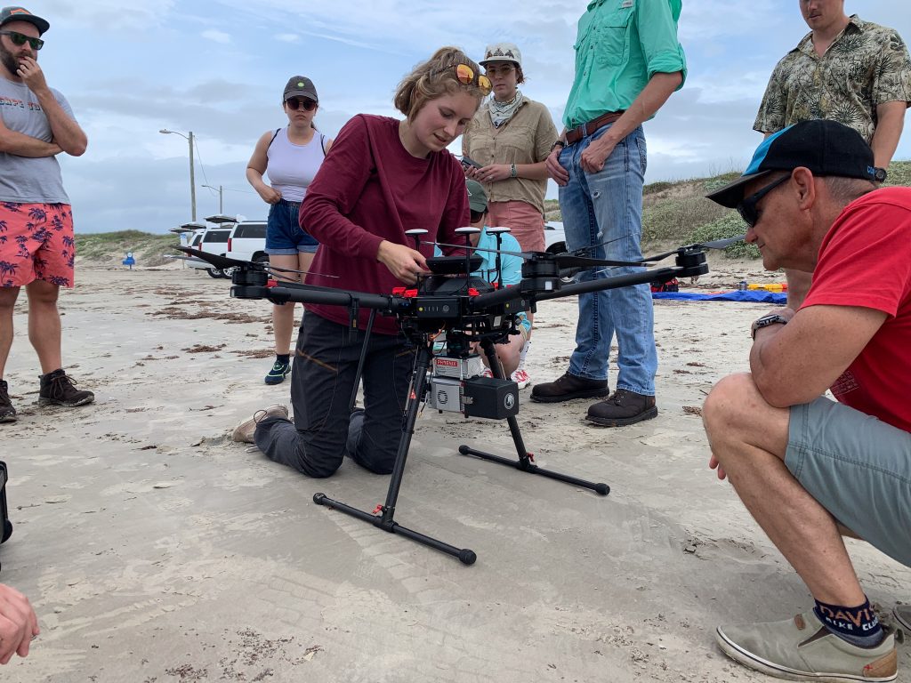 Mariel is kneeling next to the drone on the beach. People are gathered around here