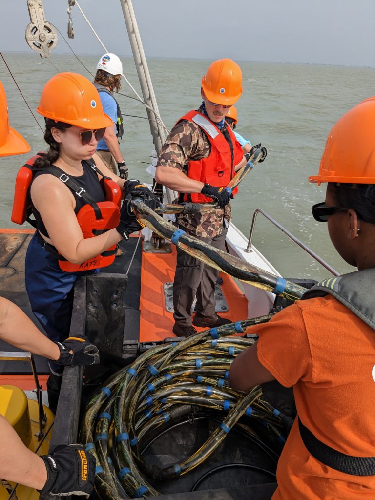 Photo of three students wearing orange hard hats uncoiling a rope-like instrument to lower into the sea.