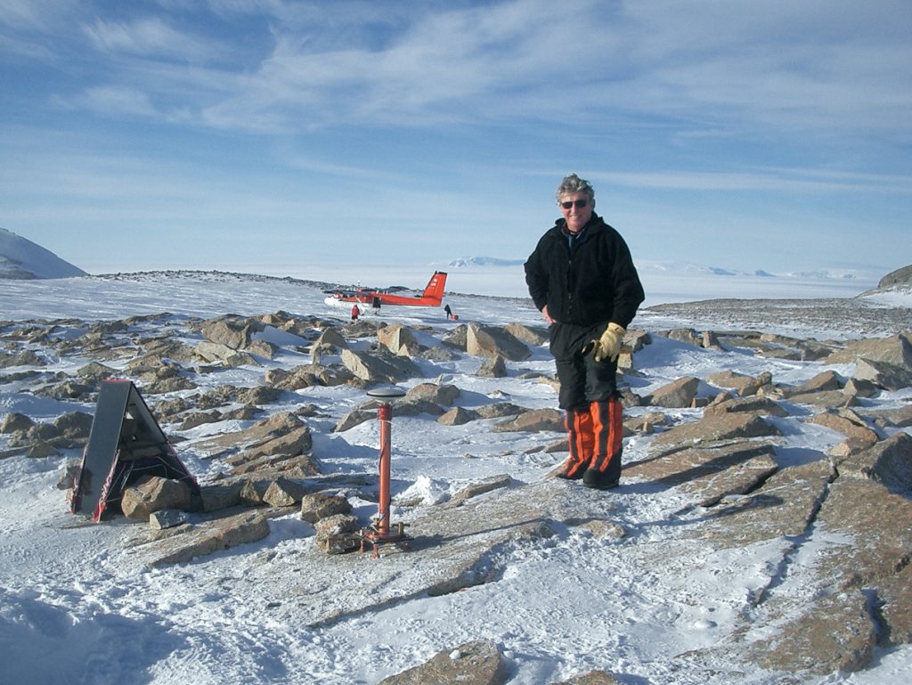 Photo of Dalziel in Antarctica. The landscape is rocky and ice covered. He is standing next to a waist-high GPS antenna. Parked behind him is a red and white Twin Otter aircraft.