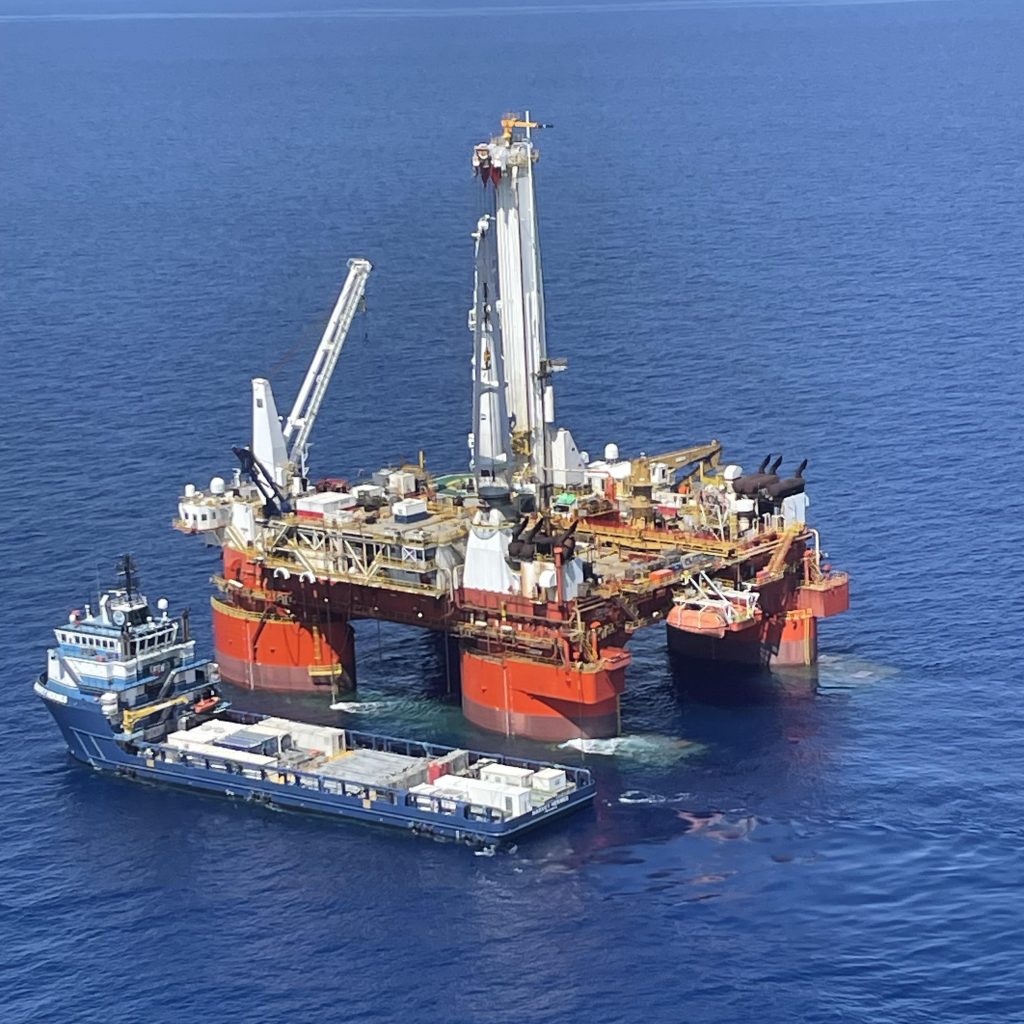 Photo of a blue freighter alongside a much larger drilling vessel surrounded by ocean. The photo is from a long distance and taken from high in the air.