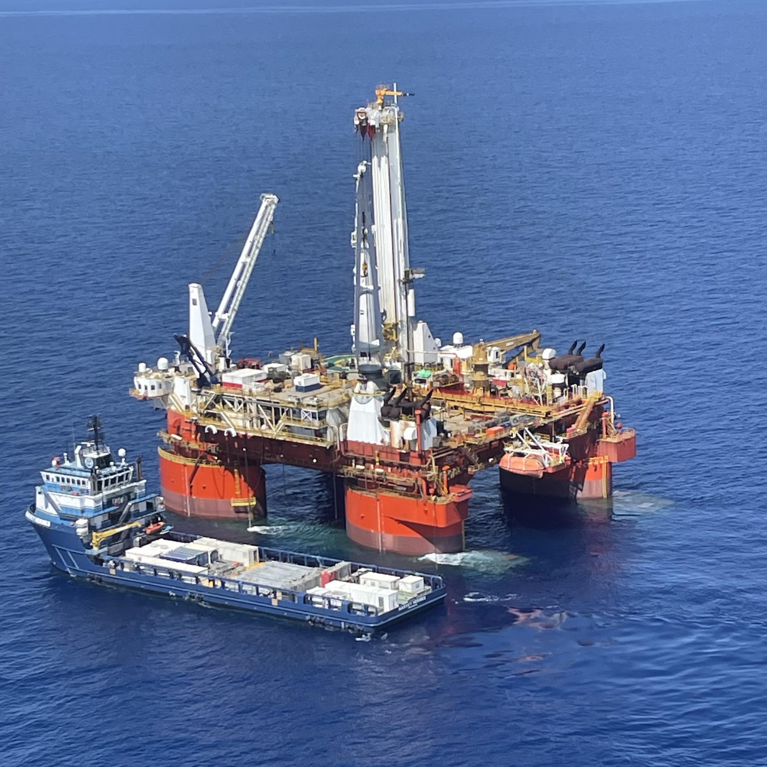 Photo of a blue freighter alongside a much larger drilling vessel surrounded by ocean. The photo is from a long distance and taken from high in the air.