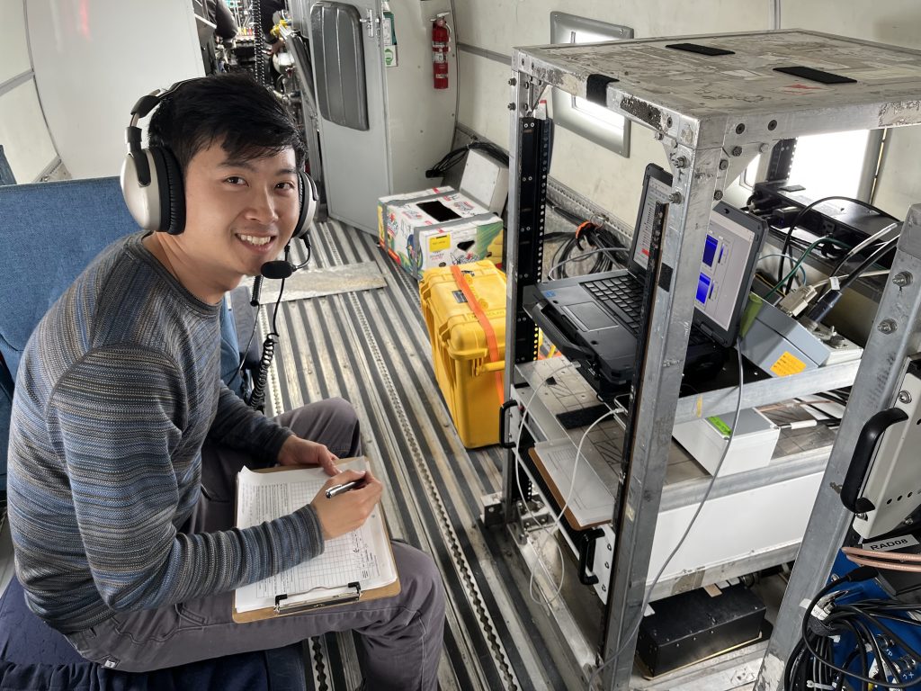 Picture of Kristian wearing headphones sitting at a control panel holding a clipboard. He is on an aircraft..