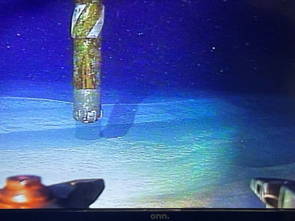 Photo of screen showing underwater camera feed from an ROV. The image shows the drill bit just above the light colored silty seafloor.