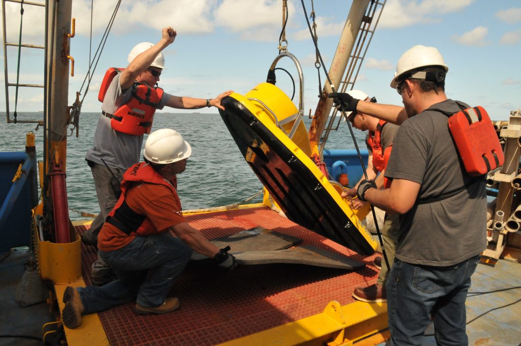Researchers wearing life vests and safety gear use pulleys and cables to lift a yellow board shaped instrument on the side of the boat.