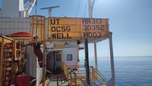 A photo of the rig flag. The flag has three lines and reads UT WRI3, OCSG 30392, WELL H003. A worker is on a service ladder next to the flag looking out over the sea.