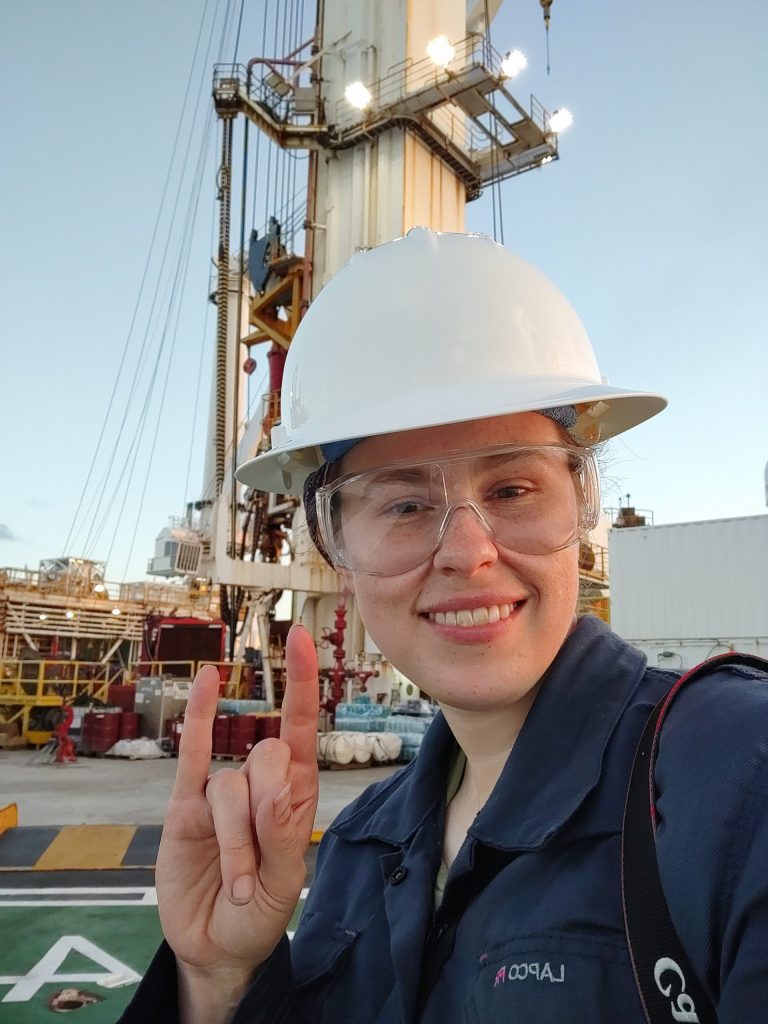 Photo of Monica on the drilling floor with cranes and machinery behind her. She is wearing protective gear and flashing a hook 'em at the camera.