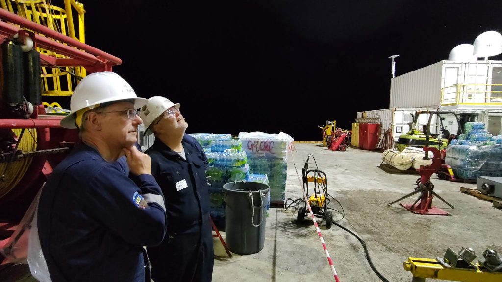 Two scientists wearing protective gear watching off-frame operations on the rig floor at night. Peter (nearest) looks pensive.
