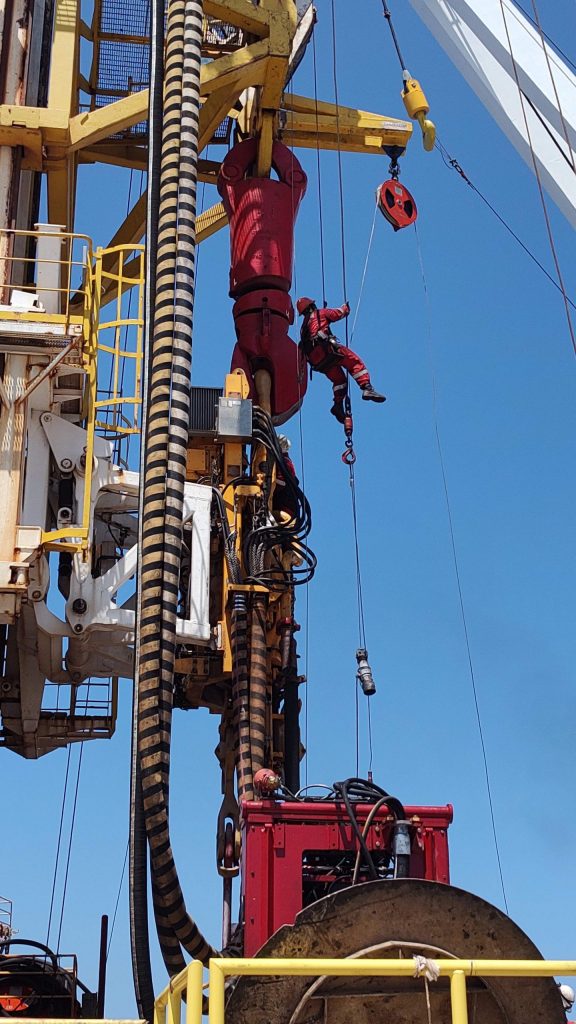 The drill worker is suspended in the air by a wire among cranes and other tall equipment. Suspended a few feet below him is a cylindrical-shaped piece of equipment about 2 feet long.