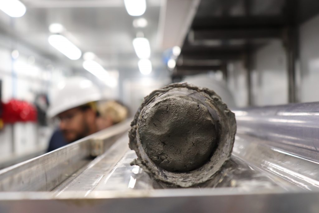 Close-up of the end of the core. The core appears like a cylinder of clay within a plastic linking - we are looking at a cross-section. Scientists are visible in the background.