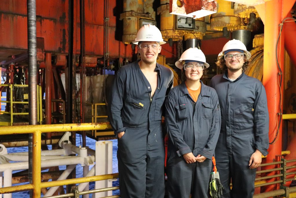 Photo of the students (and postdoc) posing for the camera in front of heavy machinery. They are wearing safety gear.