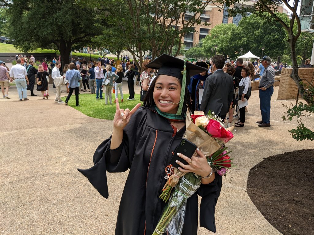 Abby flashing a hook em at the camera following her graduation. She is wearing graduation gowns and carrying flowers.