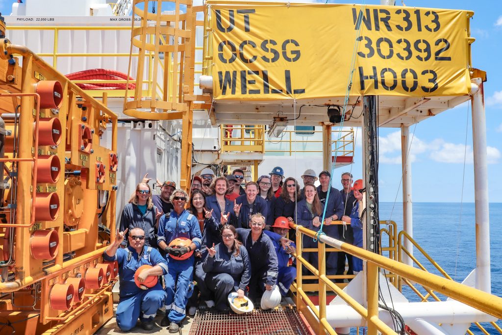 Photo of about 20 members of the science team wearing overalls and protective gear posing for a group photo under the yellow rig flag bearing the name UT and the well number. The team are flashing a hook 'em at the camera.