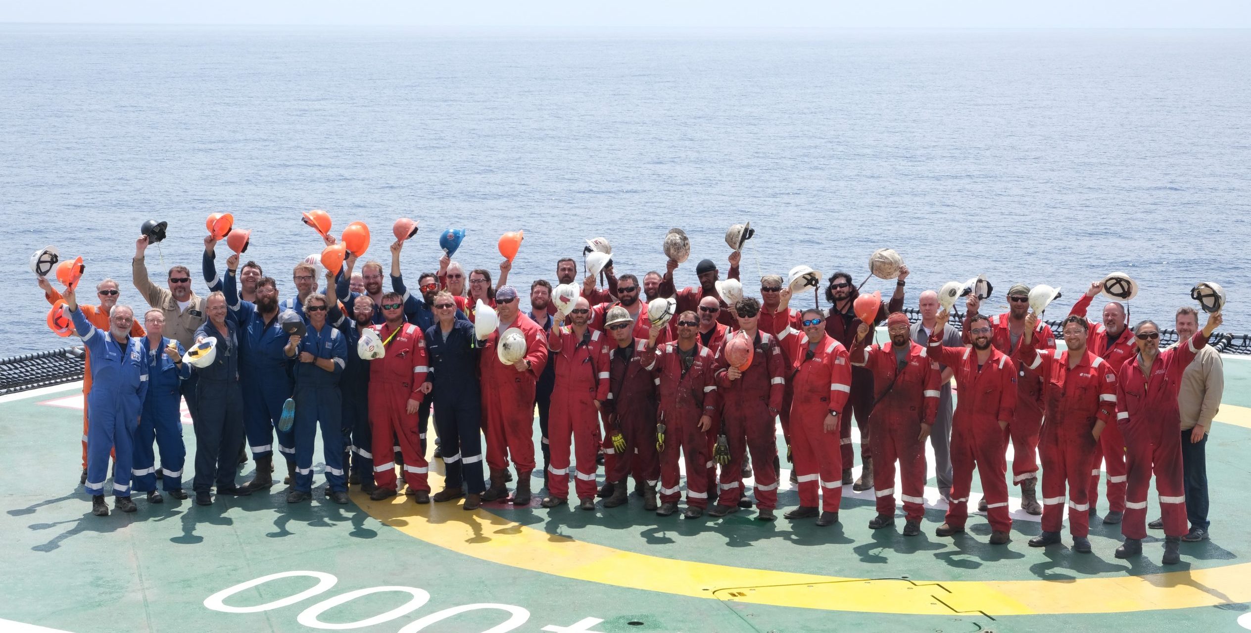 Group photo of the team standing on the helideck with the sea in the background. There are some 40 people, all wearing overalls and waving their hardhats at the camera. Words painted on the deck read "Q4000".