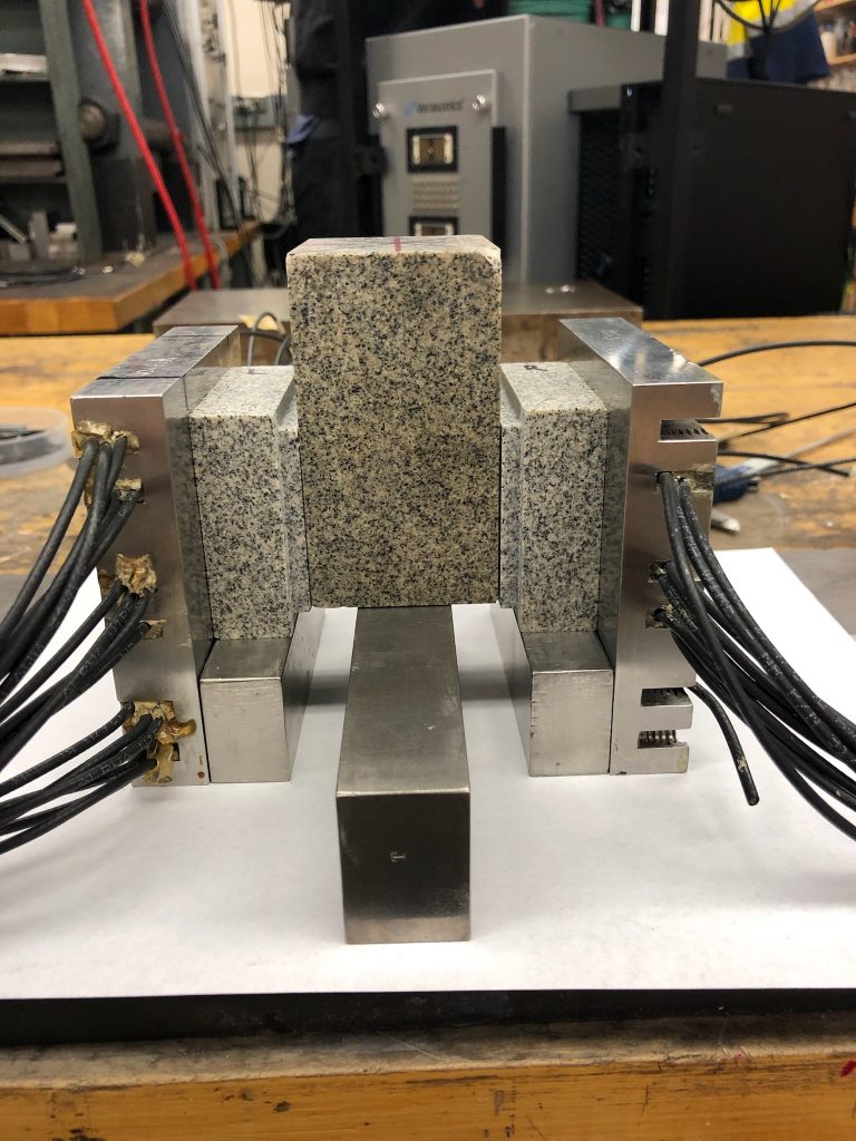 Photo of three granite-like blocks in an instrument that looks like a metal vice, connected to thick heavy cables.