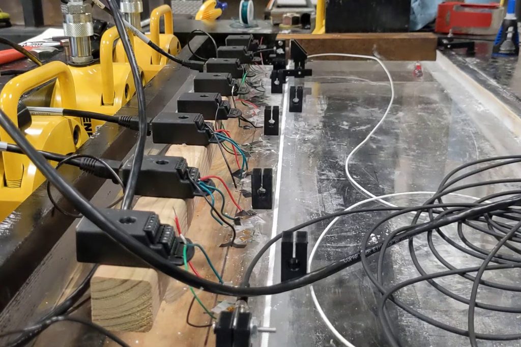 Close up photo of a line of black sensors and cables attached to a thick slab of Perspex with a white line (the fault) running up the middle. Large yellow hydraulic presses are visible to the left of the device.