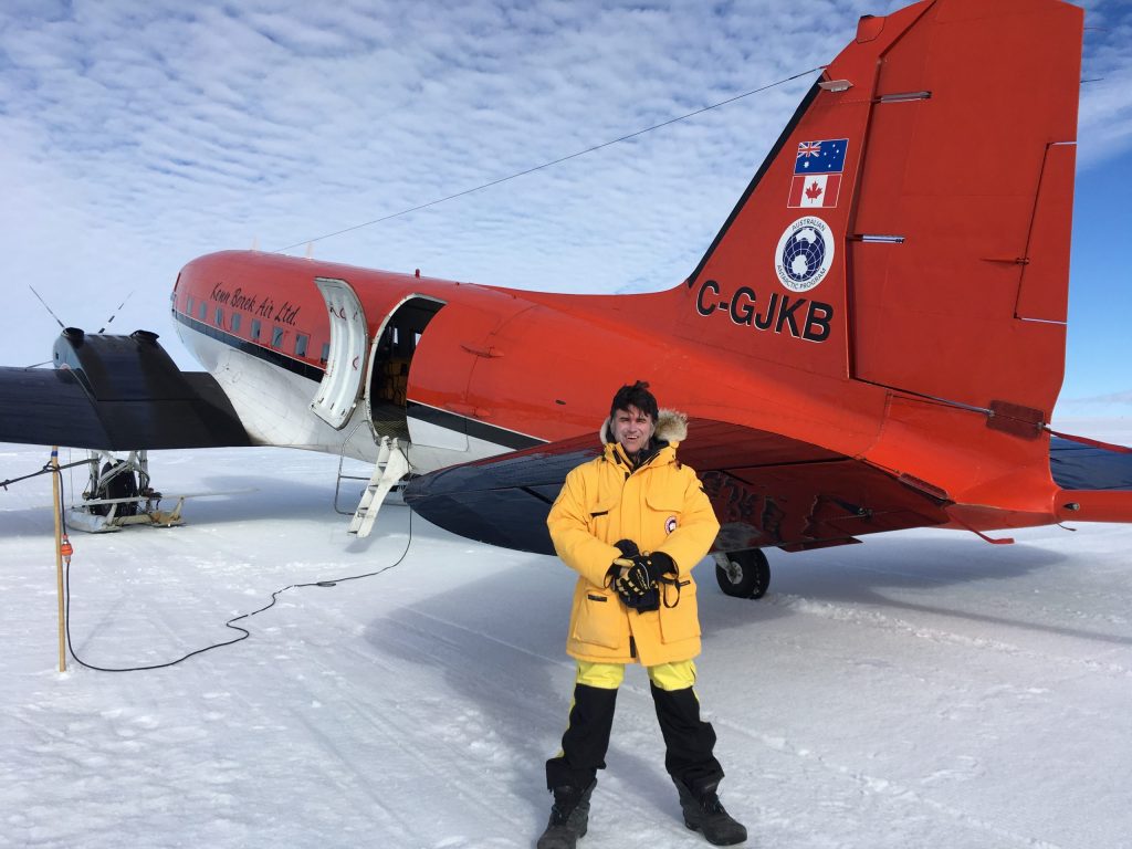 Photo of Duncan in yellow polar gear posing for a photo in front of a red DC-3 parked on the ice.