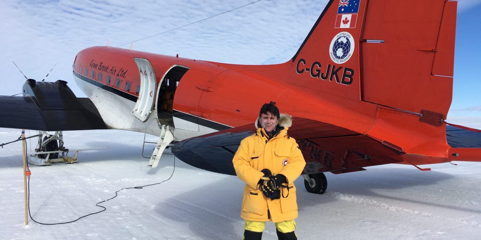 Photo of Duncan in yellow polar gear posing for a photo in front of a red DC-3 parked on the ice.