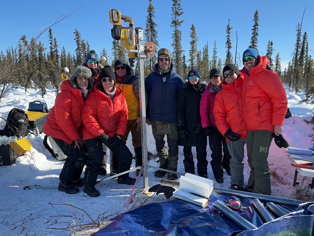 Photo of the group standing around the ice drill with snow and trees around them