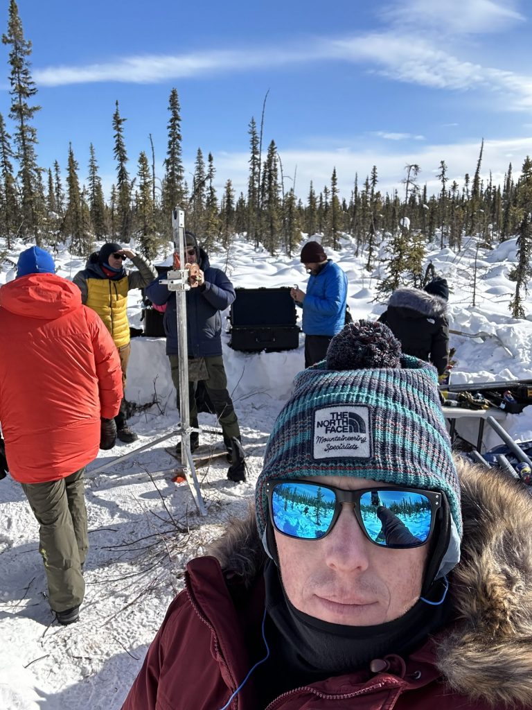 Selfie photo of Tanner in Alaska with researchers working on an ice drill behind him