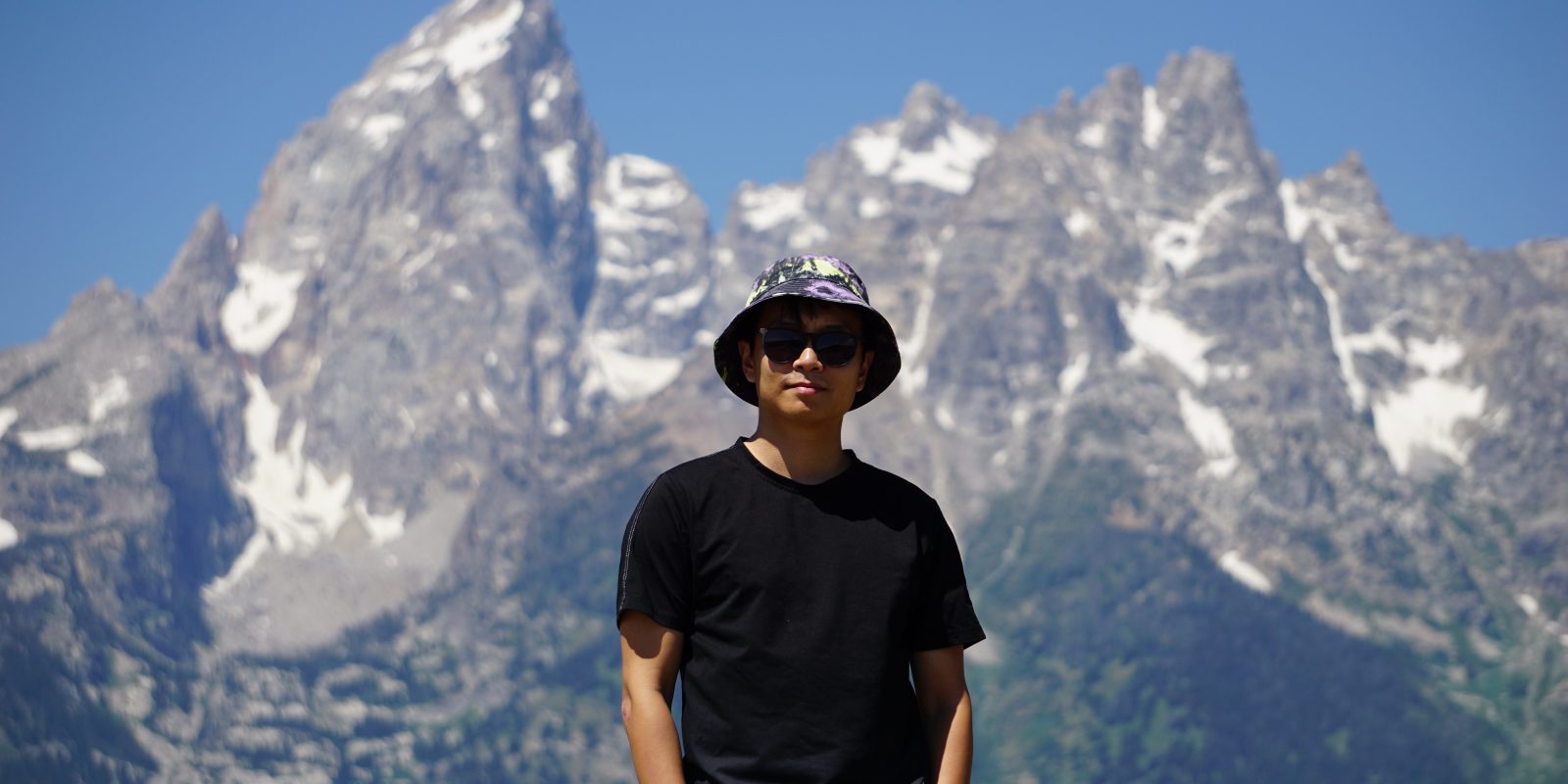 Photo of Enze wearing a hat sunglasses looking pretty chill in front of some spectacular mountains.