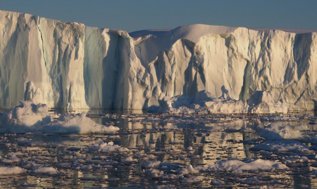 A glacial wall fills the horizon. In front the ocean is filled with ice floes and ice bergs.